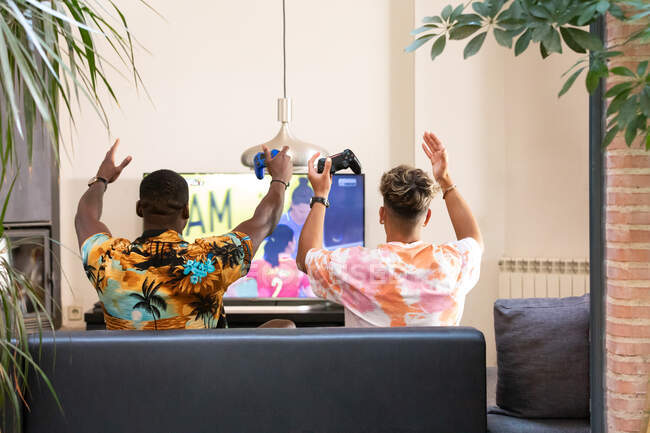 Rückansicht von anonymen multirassischen männlichen Freunden, die auf dem Sofa sitzen, während sie im Wohnzimmer ein Fußballvideospiel im Fernsehen spielen — Stockfoto