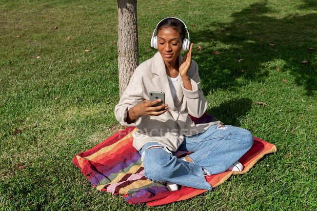 Full body of content African American female listening to music in wireless headphones while surfing cellphone on plaid near tree trunk in park — Stock Photo