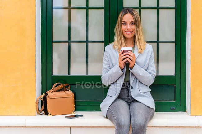 Content female in trendy outfit with paper cup of coffee looking at camera while sitting near building with purse — Stock Photo
