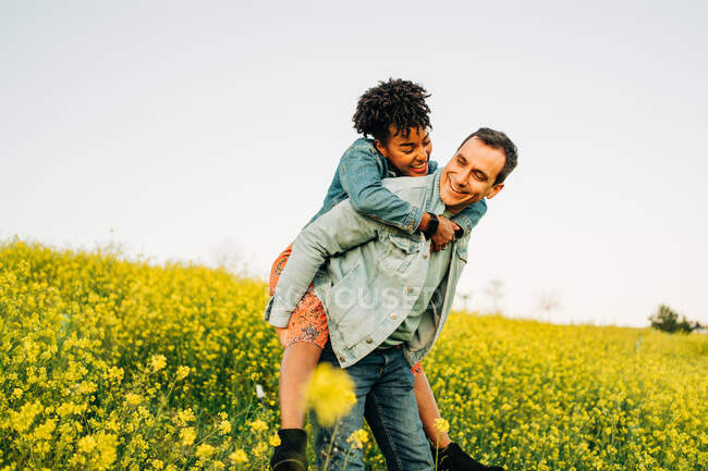 Jovem romântico sorrindo e dando passeio de piggyback para a alegre namorada afro-americana no exuberante prado amarelo florescendo no campo — Fotografia de Stock