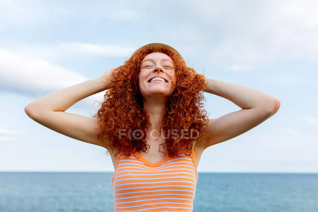 Jovem fêmea feliz com olhos fechados colocando chapéu no cabelo encaracolado na praia durante as férias — Fotografia de Stock
