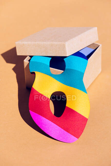 Разноцветная маска для праздничного мероприятия, помещенная в открытую картонную коробку с крышкой на оранжевом фоне в светлой современной студии — стоковое фото