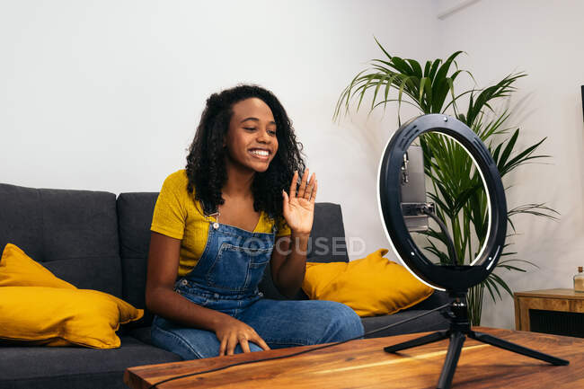 Улыбка черной женщины на диване размахивая рукой при использовании смартфона на светодиодной лампе кольцо рядом с профессиональными огнями на штативах — стоковое фото