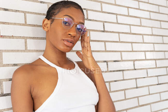 Уверенная афроамериканка с короткими волосами в стильном наряде с модными очками, стоящая на улице возле белой кирпичной стены — стоковое фото