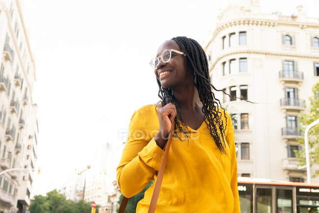Femme afro-américaine positive dans des lunettes avec les cheveux noirs debout sur la rue avec des bâtiments résidentiels en ville contre ciel sans nuages — Photo de stock