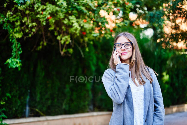 Позитивна жінка-працівниця з світлим волоссям стоїть на тротуарі і розмовляє по мобільному телефону, дивлячись у далечінь. — стокове фото