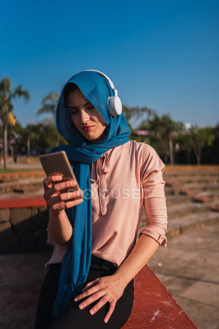 Mujer árabe en pañuelo para la cabeza y auriculares navegando por el teléfono móvil y escuchando música mientras disfruta de un día soleado en la ciudad - foto de stock