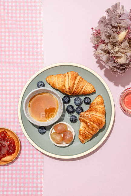 Vista superior de deliciosos croissants servidos em prato cerâmico com mirtilos frescos e geléia colocada na mesa rosa — Fotografia de Stock