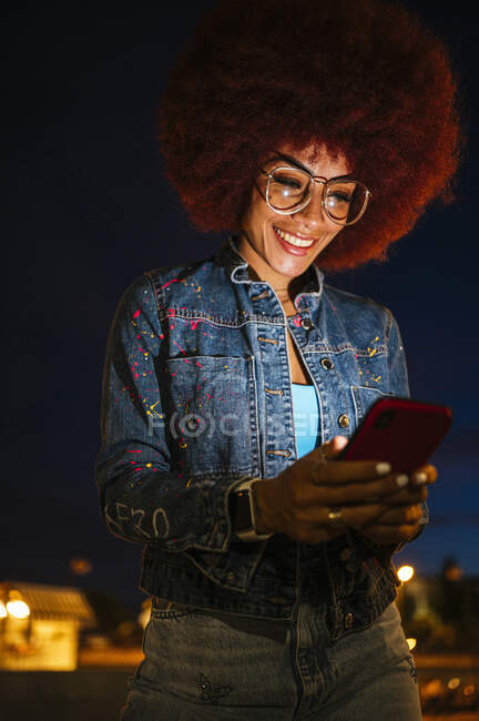 From below positive female with Afro hairstyle and modern clothes text messaging on cellphone while walking around the city at night — Stock Photo