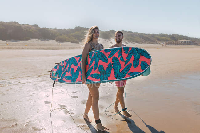 Cuerpo completo de pareja deportiva en traje de baño con tablas de surf mientras está de pie en la orilla del mar de arena en el resort tropical - foto de stock