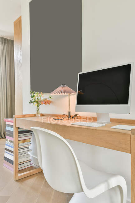 Ordenador moderno con pantalla negra y teclado colocado en escritorio de madera con lámpara y florero en sala de trabajo ligera con interior minimalista - foto de stock