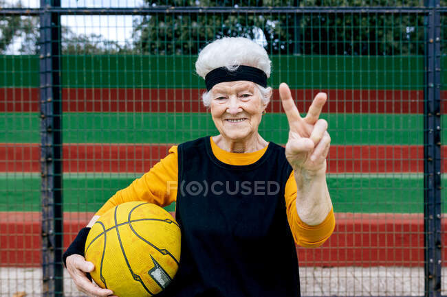 Mujer madura optimista en ropa deportiva y diadema mirando a la cámara mientras está de pie en la cancha pública de baloncesto con pelota durante el entrenamiento - foto de stock