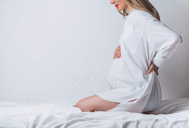 Vista laterale di anonimo giovane incinta dolce femmina toccando pancia mentre seduto sul letto — Foto stock