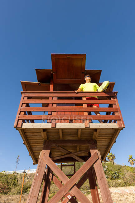 Low angle of brave lifeguard in sunglasses standing on wooden rescue tower and monitoring safety under blue sky — Stock Photo