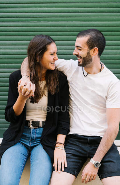 Fröhlich verliebtes junges hispanisches Paar in lässiger Kleidung lacht und schaut einander an, während es sich in der Nähe der grünen Mauer auf der Stadtstraße umarmt — Stockfoto