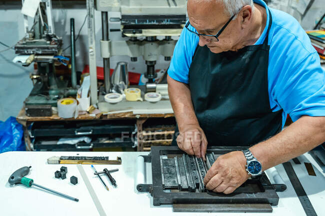 Alto ângulo de cultura grave idoso artesão masculino em avental e óculos preparando armação metálica para impressão de tipografia em estúdio — Fotografia de Stock