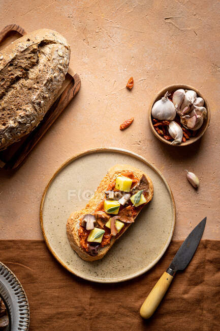 Вид сверху на тост с кубиками сыра и слайсами грибов, подаваемых на тарелке рядом с буханкой свежего хлеба и миской чеснока на кухне — стоковое фото