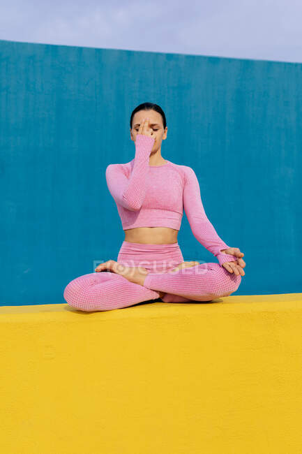 Мирна жінка в рожевому активаторі сидить в Падмасані з руками Намасте і медитує під час сеансу йоги на синьому і жовтому фоні. — стокове фото