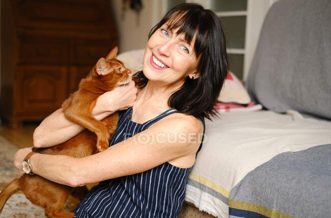 Хозяйка, сидящая на диване и играющая с домашней короткошерстной абиссинской кошкой дома — стоковое фото