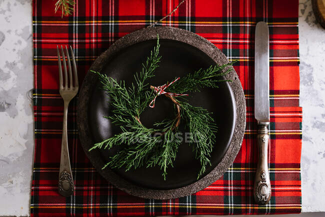 Зверху Різдвяний стіл з вінком на керамічній тарілці з столовими приборами на червоній картатій скатертині на фоні — стокове фото