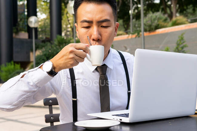 Wistful young Asian male entrepreneur with cup of hot drink and netbook in urban cafeteria table in daylight — Stock Photo