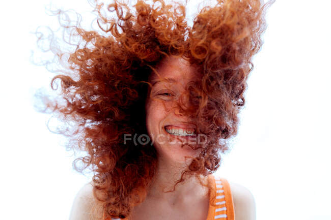 Desde abajo encantado joven mujer cubriendo la cara con el pelo rojo rizado mientras se ríe felizmente mirando la cámara - foto de stock