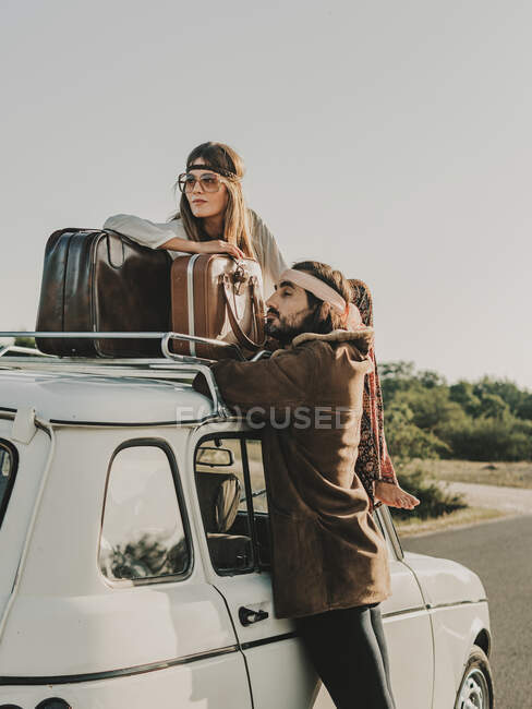 Vista lateral de pareja hippie romántica mirando hacia otro lado mientras está sentado en el automóvil viejo temporizador con maleta durante el viaje en la naturaleza - foto de stock