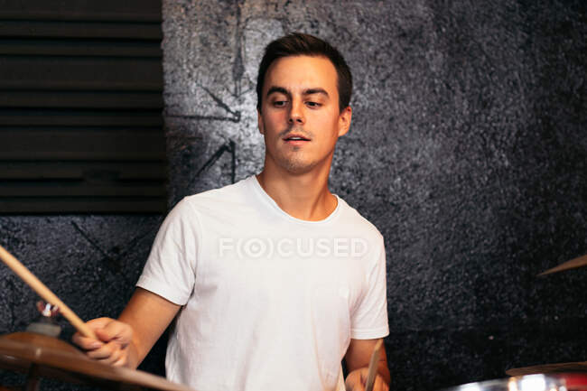 Концентрированный молодой музыкант в повседневной одежде играет на тарелке в ярком клубе рядом с серой стеной — стоковое фото