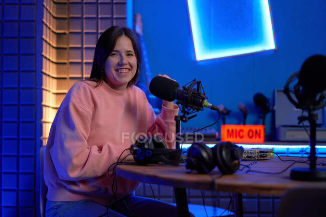 Fröhliche junge Radiomoderatorin lächelt und blickt in die Kamera, während sie während der Podcast-Aufzeichnung im dunklen Studio mit Neonlicht am Tisch mit Mikrofonen und Kopfhörern sitzt — Stockfoto