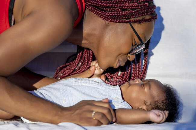 Vista lateral de la madre afroamericana amante de las cosechas con trenzas rojas en gafas que tocan y miran al bebé dormido en un día soleado - foto de stock
