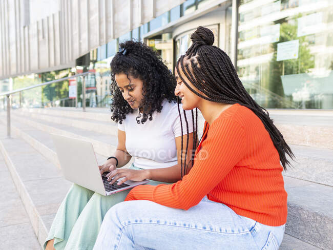 Cheerful diverse female friends in trendy clothes sitting on stair and browsing laptop on city street in summer day — Stock Photo