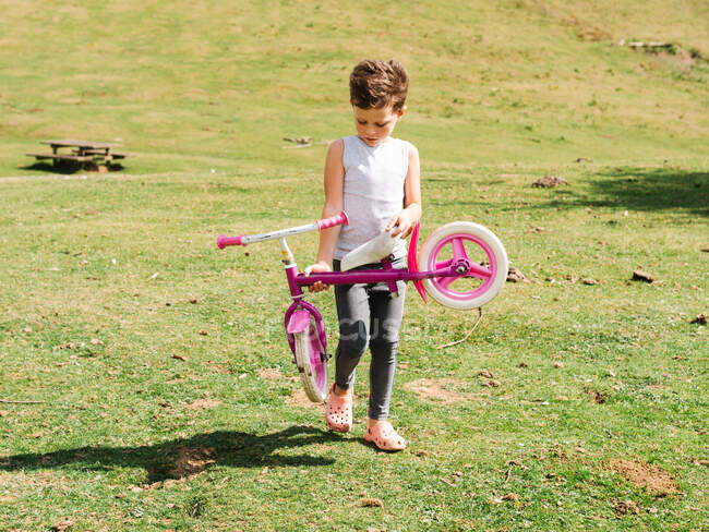 Cuerpo completo de chico serio con bicicleta rosa paseando en el campo de hierba cerca de la colina verde en la naturaleza en el día de verano - foto de stock
