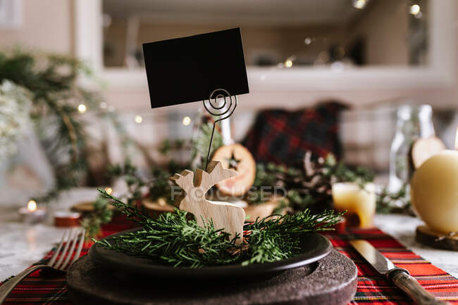 Різдвяний стіл з оленями дерева з порожньою карткою на керамічній тарілці на червоній картатій скатертині на фоні — стокове фото