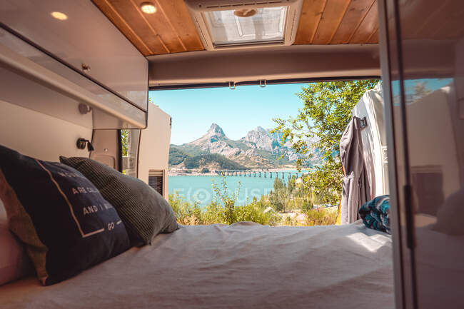 Comfortable bed with cushions and blanket in cozy traveling caravan parked in mountains near blue lake in Riano — Stock Photo