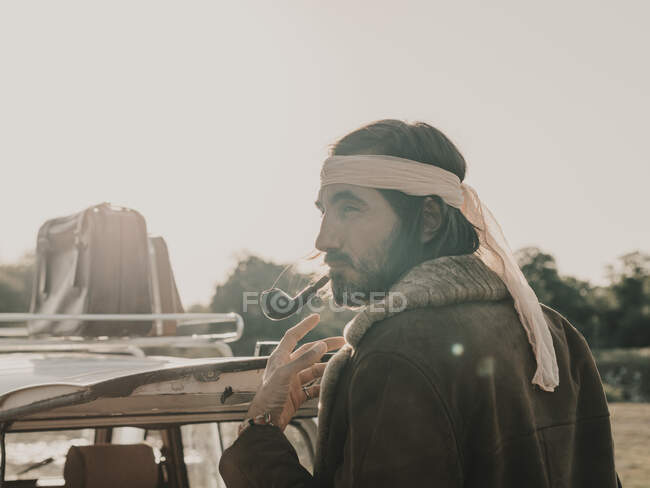 Side view of unshaven male hippie in headscarf smoking pipe while standing near old timer car with suicase during trip in nature — Stock Photo