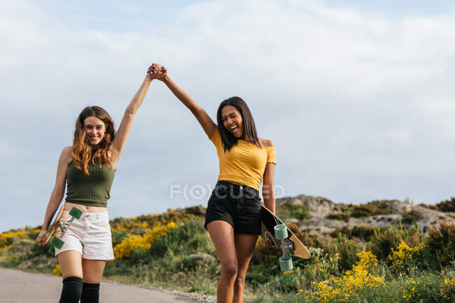 Felices novias multiétnicas cogidas de la mano mientras caminan por la carretera con longboards - foto de stock