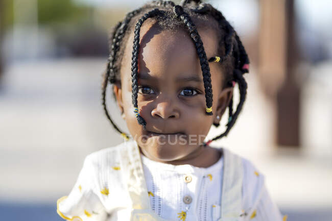 Афроамериканская маленькая девочка с косичками в стильной одежде, стоящая на улице напротив здания в солнечный день — стоковое фото
