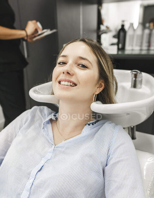 Positive junge Dame in lässiger Kleidung, die in die Kamera schaut, während sie in der Nähe des Waschbeckens sitzt und sich darauf vorbereitet, sich im Friseursalon die Haare zu waschen — Stockfoto