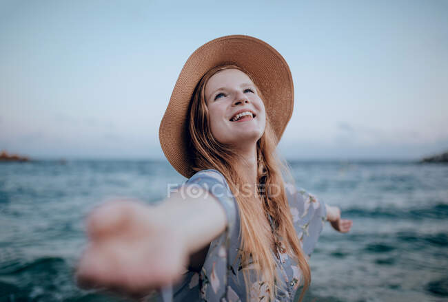 Улыбающаяся молодая женщина в повседневной одежде, стоящая на песчаном пляже рядом с волнистым морем, с распростертыми объятиями летом — стоковое фото