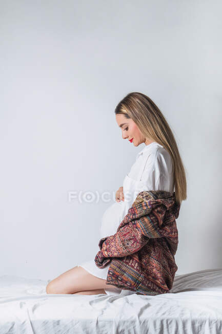 Vista laterale della giovane dolce femmina incinta che tocca la pancia mentre è seduta sul letto — Foto stock
