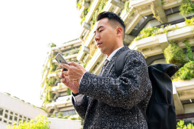 Von unten schaut ein junger männlicher Unternehmer mit Krawatte auf den Bildschirm, während er in der Stadt mit dem Handy spricht — Stockfoto