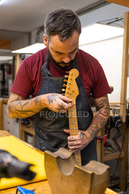 Seriöser männlicher Meister in Schürze steht neben Tisch mit unterschiedlichem Zubehör und repariert E-Gitarre in Lichtwerkstatt — Stockfoto