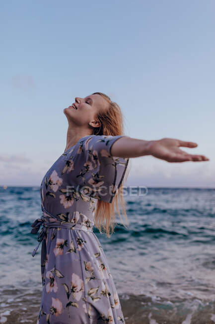Delighted female with long hair in trendy dress standing on seashore in summer evening — Stock Photo