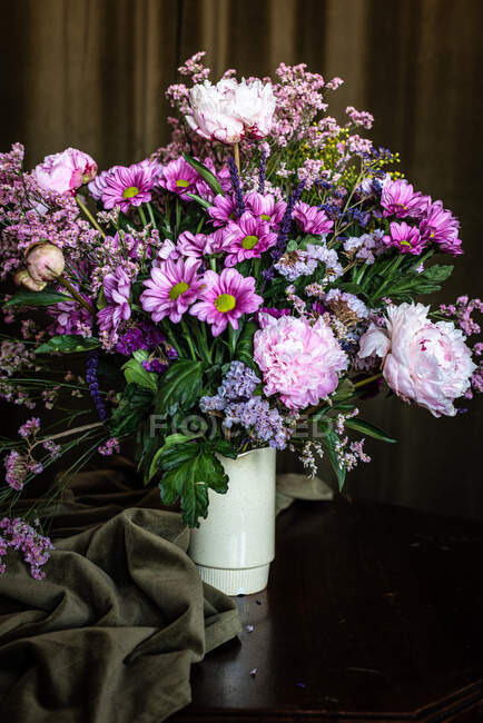 Bouquet de pivoines fraîches colorées et de chrysanthèmes dans un vase blanc posé sur une table en bois dans une pièce sombre — Photo de stock