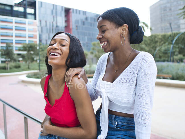 Delighted African American female friends in stylish outfits with eyes closed while leaning on metal railing near contemporary building in city — Stock Photo