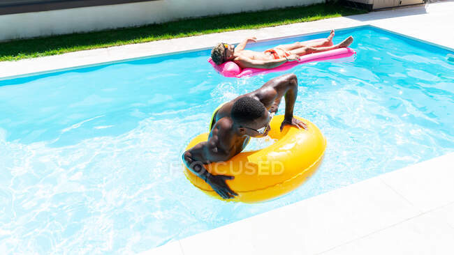 Amis masculins multiraciaux en short de bain couché sur anneau gonflable et matelas tout en bronzant dans la piscine le jour ensoleillé d'été — Photo de stock