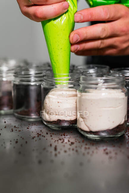 Crop anonymous male confectioner pouring yummy cream from pastry bag into glass jar while preparing appetizing layer dessert in kitchen — Stock Photo