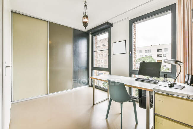 Chaise au bureau avec ordinateur moderne et lampe placée dans une pièce lumineuse spacieuse avec fenêtre et placard dans l'appartement — Photo de stock