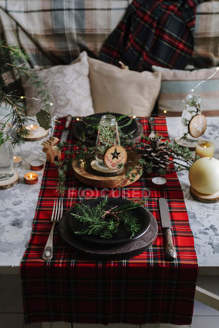 Ajuste de mesa de Navidad con corona en el plato, adornos decorativos de madera y mantel a cuadros rojo con luces amarillas en el fondo - foto de stock