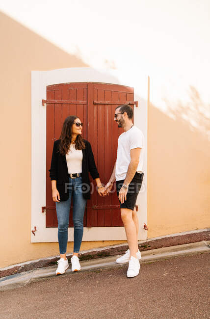 Positive young ethnic couple in casual clothes holding hands on aged stone building in city park on sunny day — Stock Photo